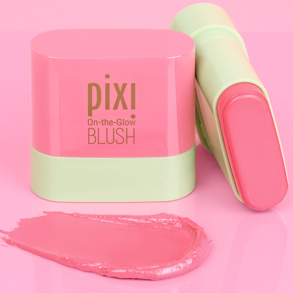Pixi On-the-Glow Blush Petite stick shown with its pink and mint green cap on, alongside a smear of the pink cream blush on a solid pink background.