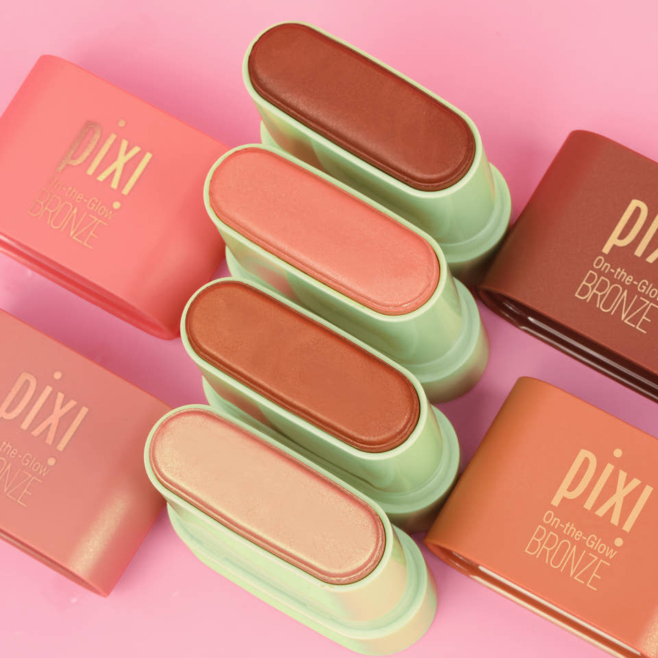 Six Pixi On-the-Glow Bronze face powders in various warm shades, arranged vertically in the center with their coral-pink and terracotta-colored rectangular boxes on a solid pink background. view 2