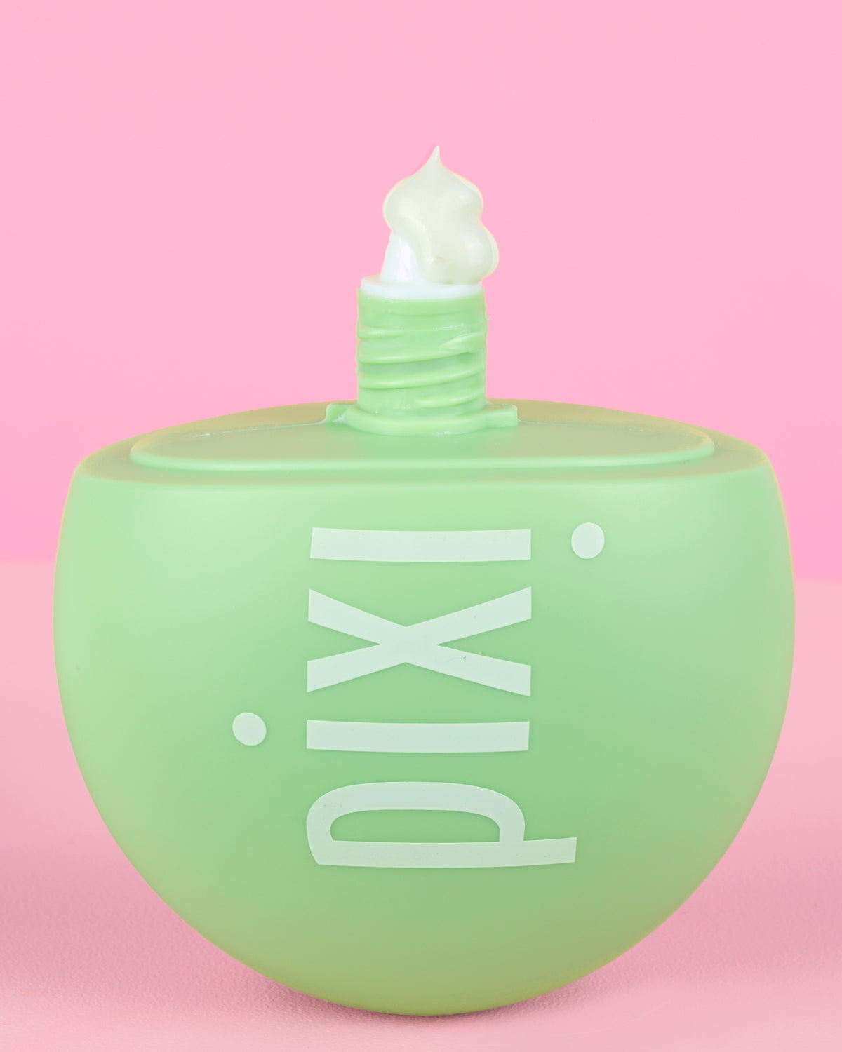Pixi skincare HandHero bottle in a light mint green color with white lettering, shown with white cream dispensed from the top against a solid pink background.