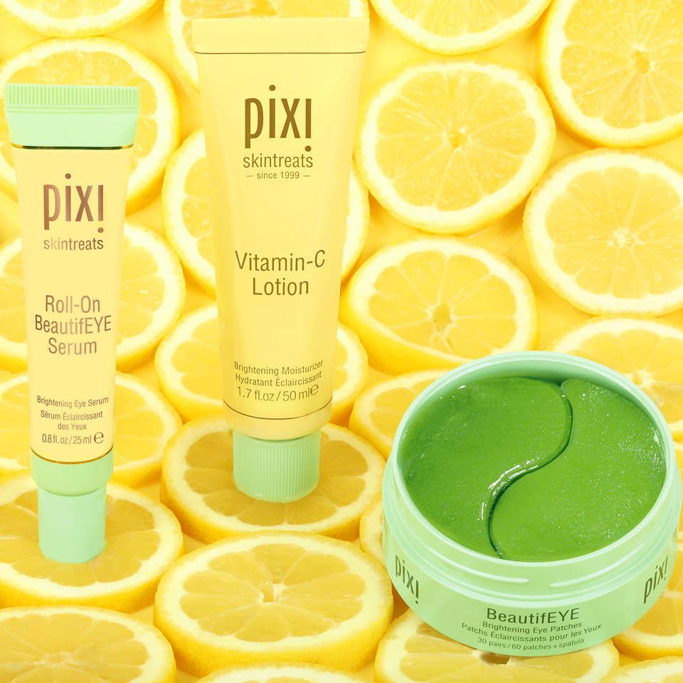 Three Pixi Skintreats BeautifEYE products arranged on a background of sliced lemons: a Roll-On BeautifEYE Serum tube, a Vitamin-C Lotion tube, and an open BeautifEYE jar containing bright green eye patches. The products use yellow and mint-green packaging. view 2